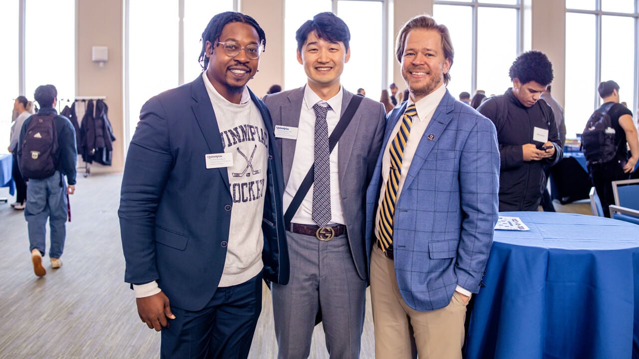 Three male students standing together and smiling in the foreground of a busy event space.