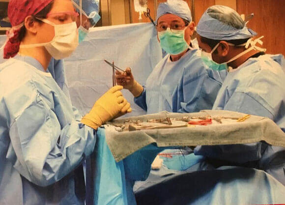 Alex Gerity, left, stands in blue scrubs and a face mask holding instruments across an operating table from two men in scrubs and face masks performing surgery.