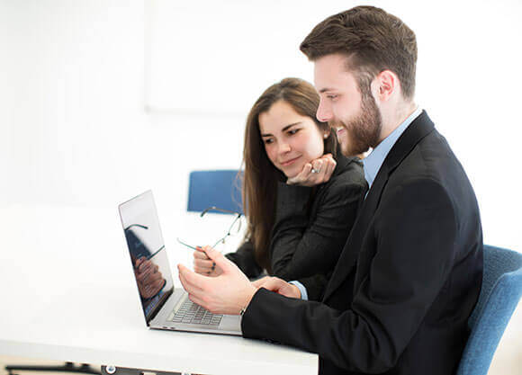 A male and female student in business attire sit at a table looking at an open laptop.