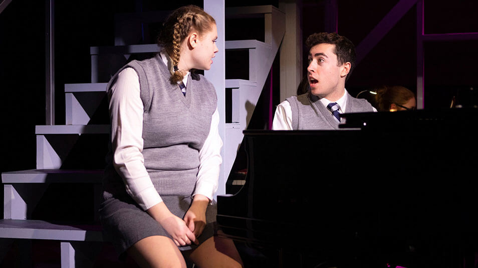 Two students sit side-by-side and sing a song together during a theater scene