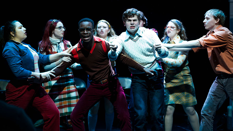 A group of student actors surround the two main characters who look frightened in a theater scene