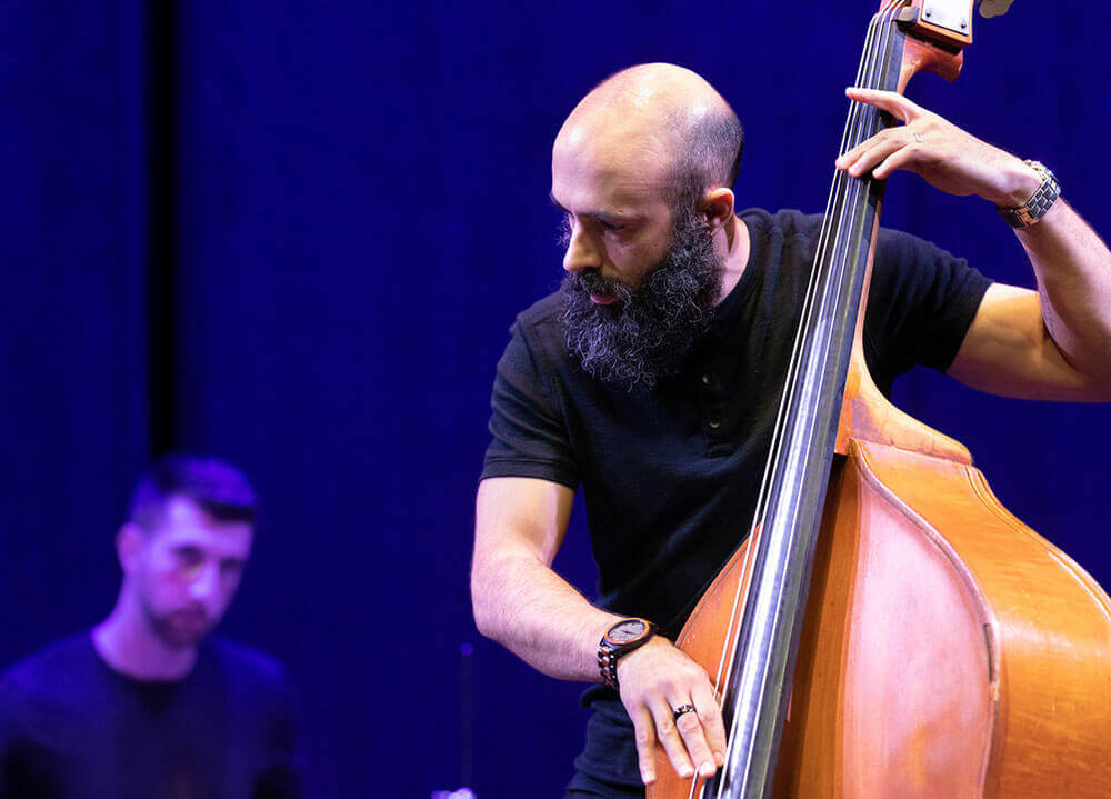 A student plucks the strings of an upright bass with an out of focus drummer in the background