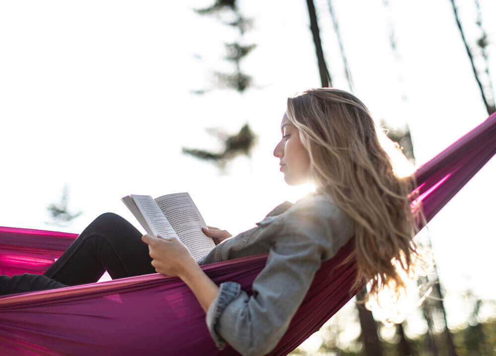 A student lays in a purple hammock reading a book
