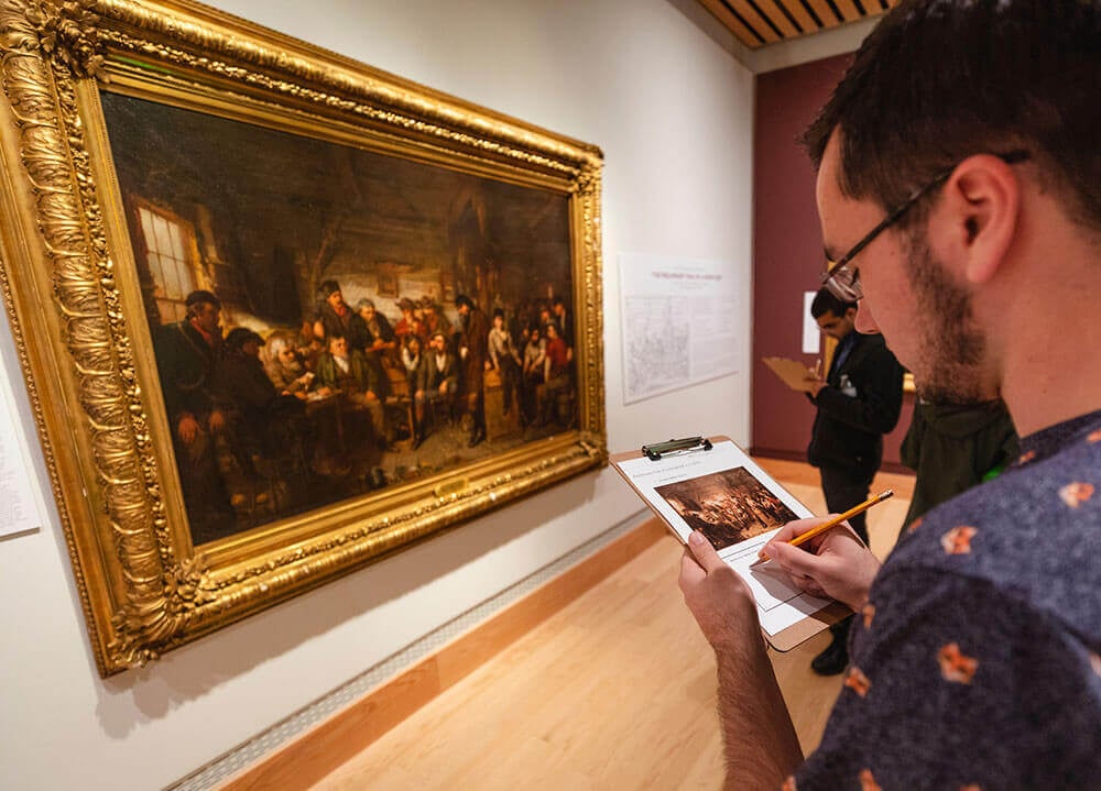 A student takes notes on a piece of paper while studying a large painting in a museum