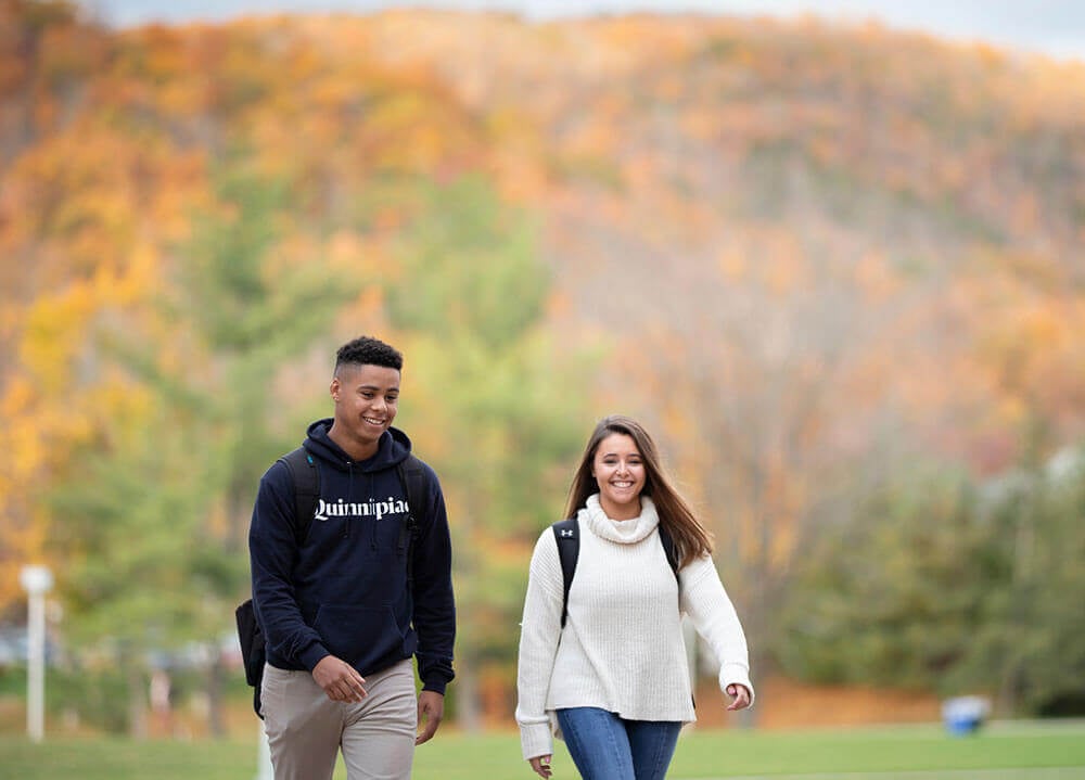 Two Quinnipiac students walk the Mount Carmel Campus quad with fall foliage in the background