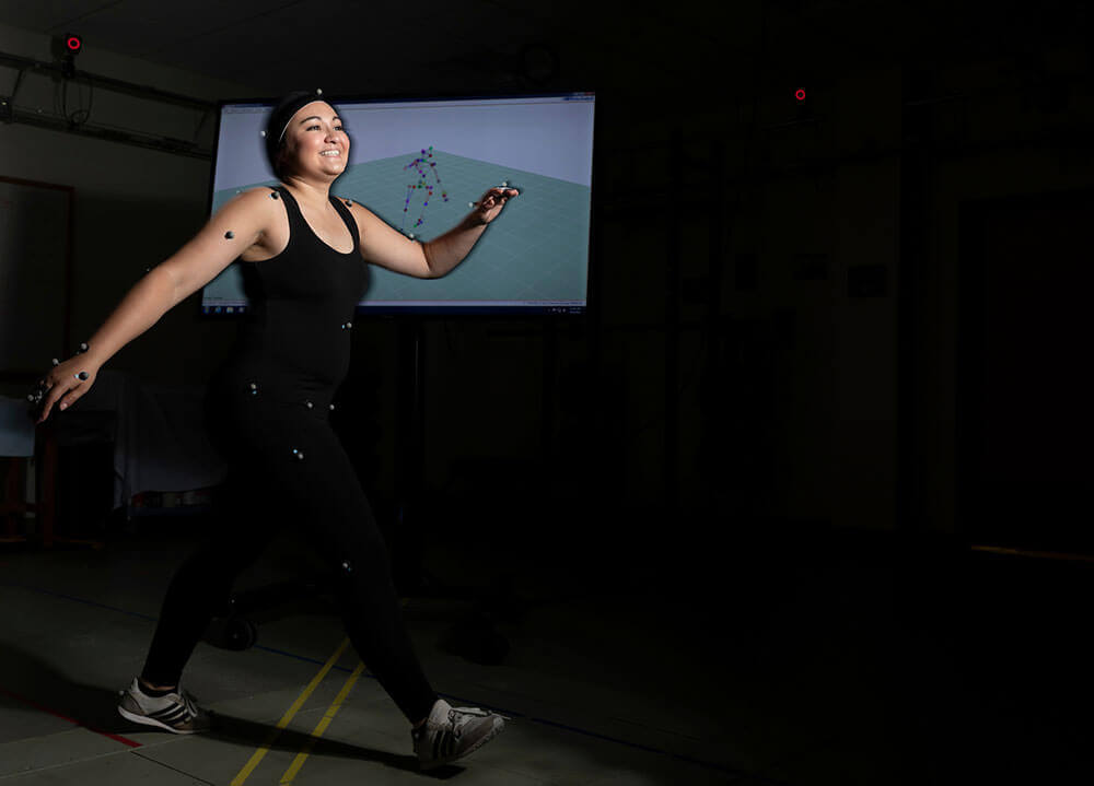 A game design student wears a black motion tracking suit with a screen showing her digital character in the background