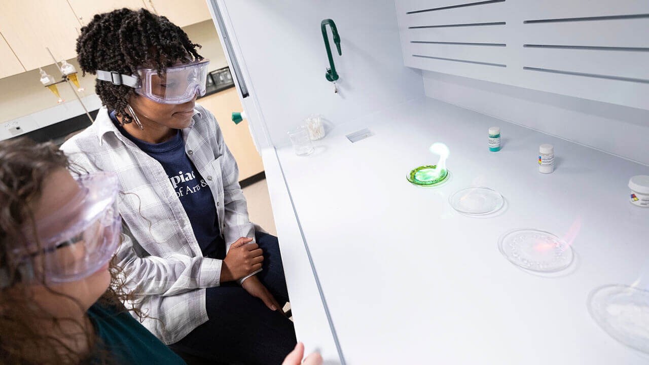A chemistry student and professor wearing goggles observe a green flame in a lab clasroom