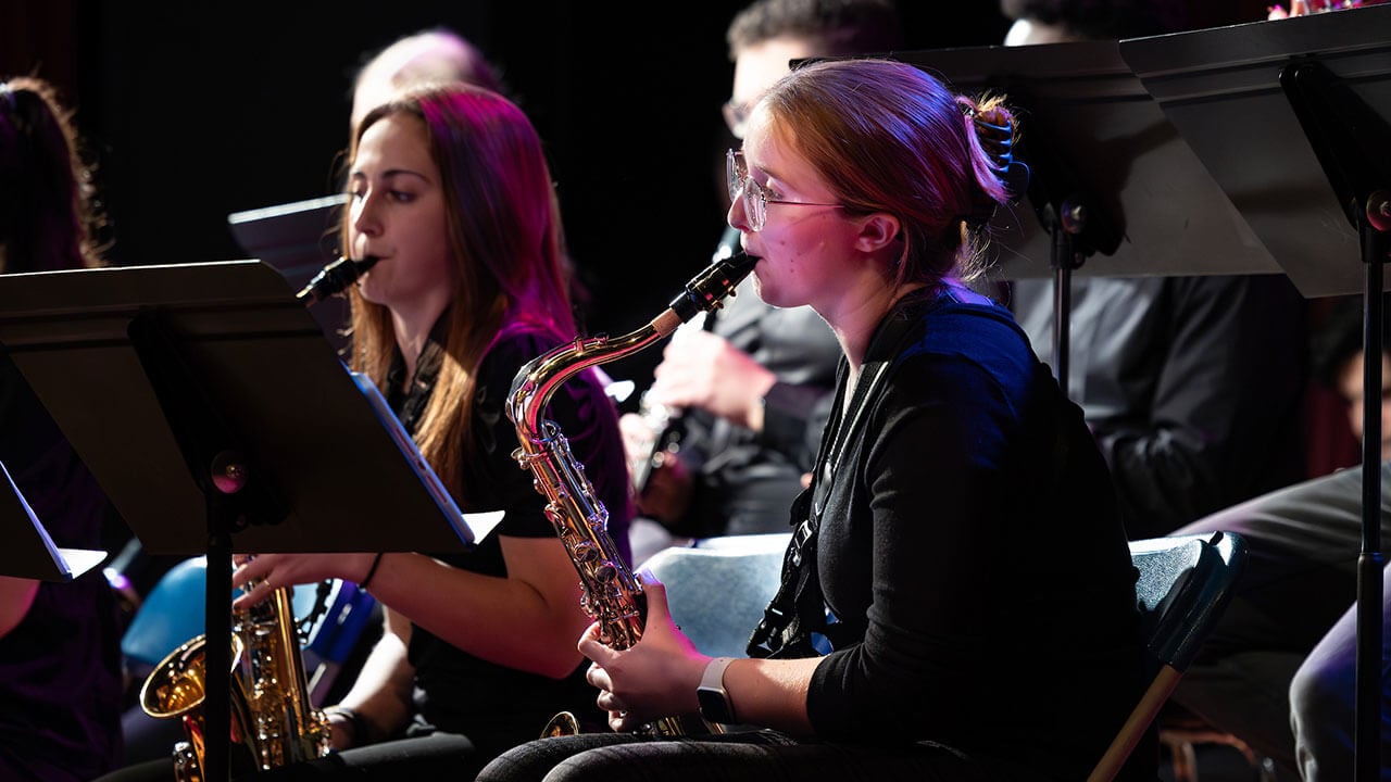 A student with glasses plays their saxophone at their music stand during the winter concert.