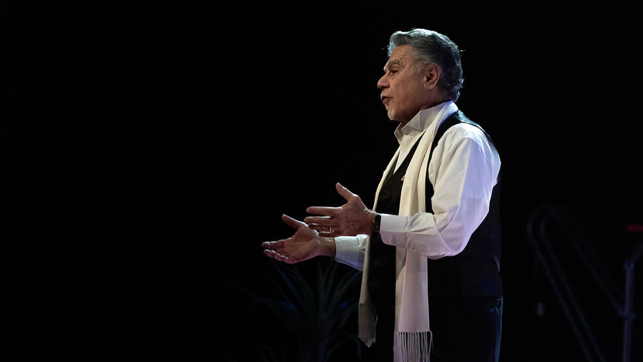 An individual wearing a black vest and white scarf speaks to the audience during a winter concert.