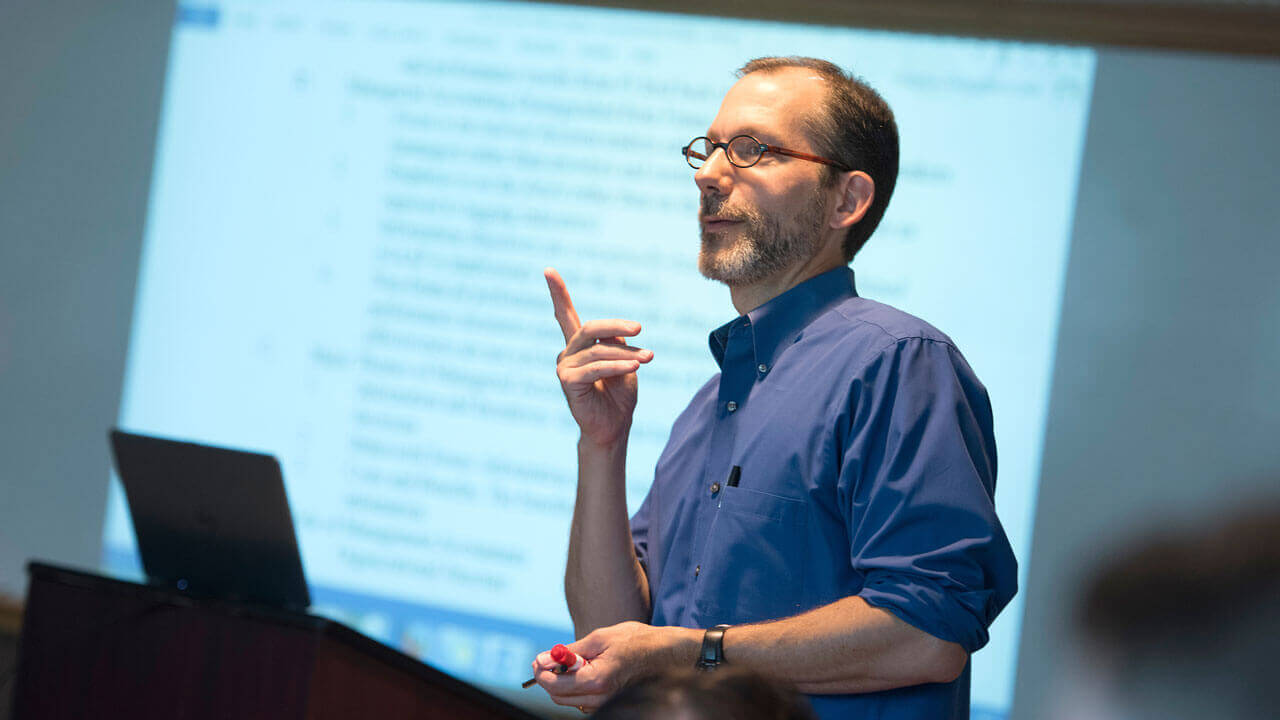 Professor Chris Hodgdon gestures his hand as he lectures in front of a screen