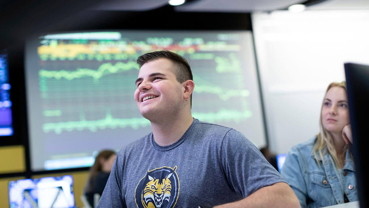 A student smiles with a large monitor displaying stock market figures in the background