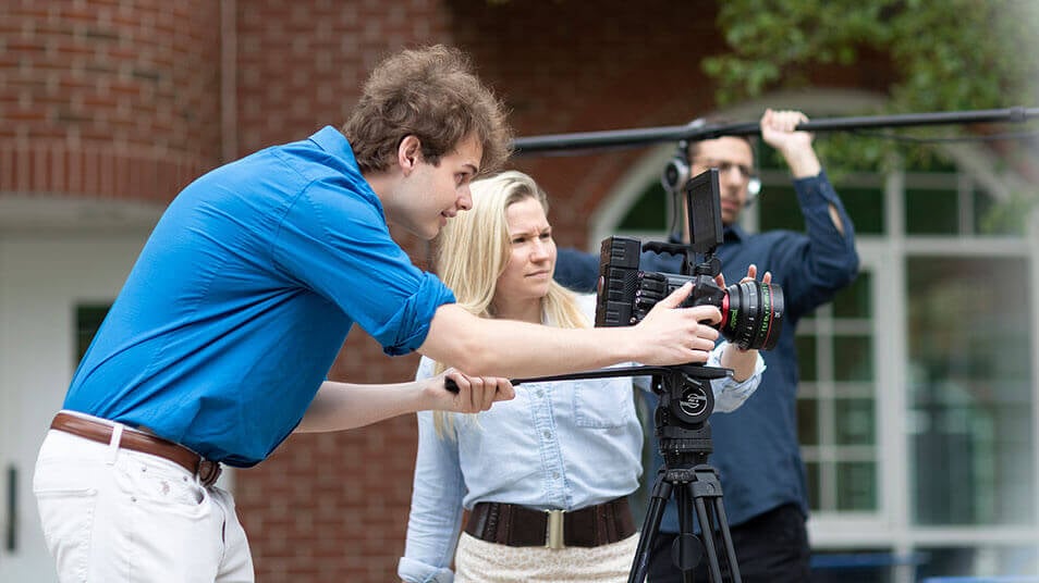 A student lines up his film camera outside on the quad while another student holds a boom mic in the background