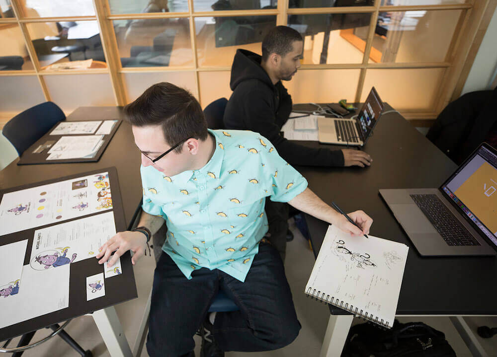 A graphic design student examines his printed portfolio laid out on a desk