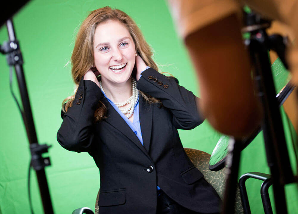 A communications student dressed in a suit gets prepared to take a professional headshot with a green screen behind her