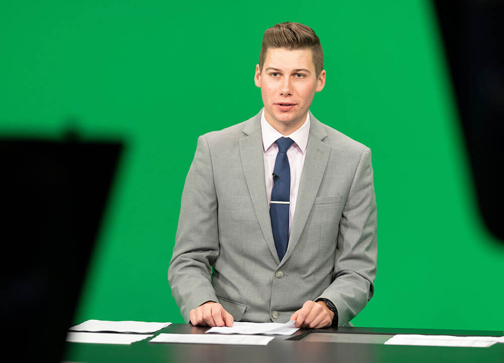 A journalism student wearing a suit and tie sits at the news desk in our communications center