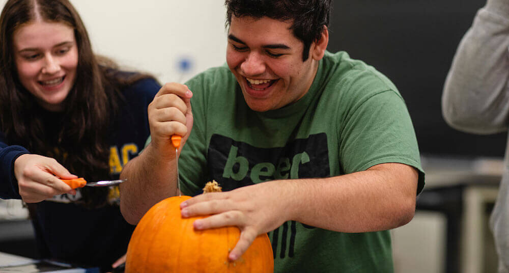 An engineering student uses a knife to carve the top off of a pumpkin