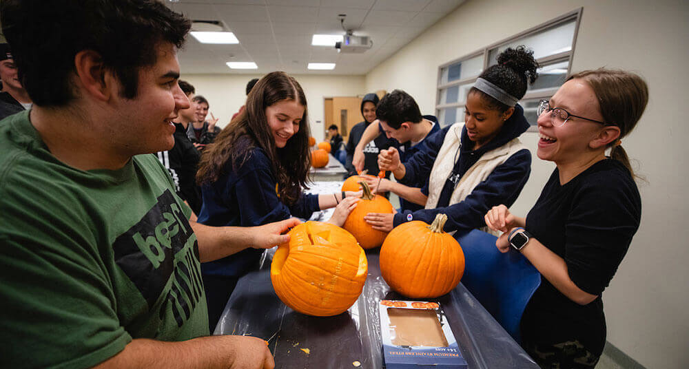 Engineering students laugh and smile as they carve designs on their pumpkins