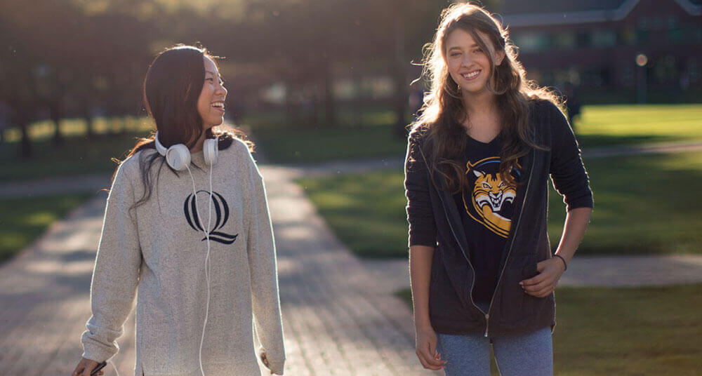 Two students wearing Quinnipiac shirts walk on the quad in the fall