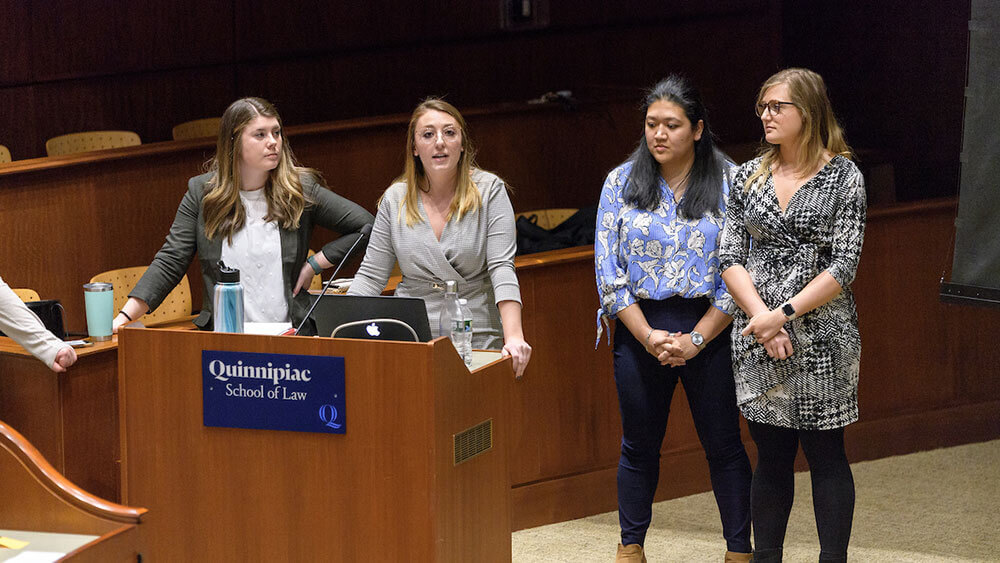 Four law students stand at the podium in the ceremonial court room during a conference