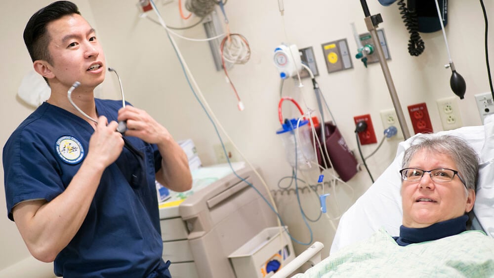 Medical student Kevin Kuo puts a stethoscope on while checking on a patient in a hospital bed