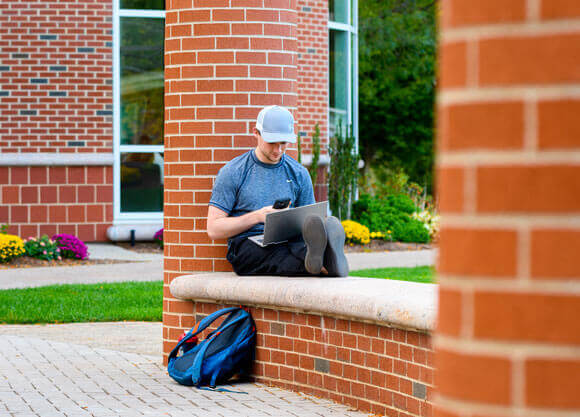 A student sits outside using his laptop and cell phone to check his admissions status