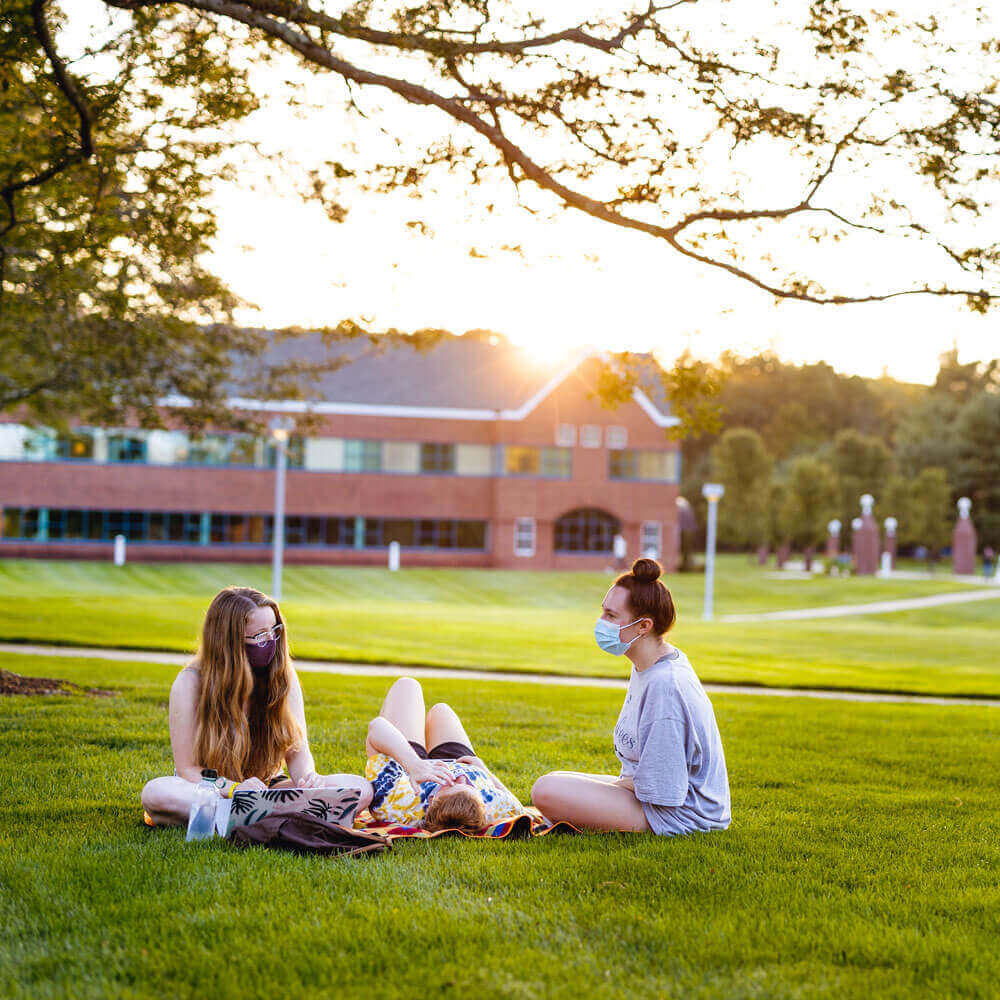 A group of students wearing masks lounging on the grass on the Quinnipiac Quad