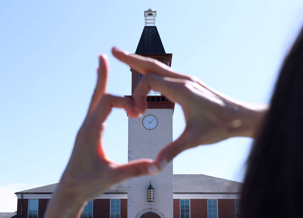 A student forms her hand into a heart with the Quinnipiac clocktower in the background