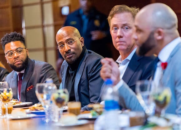 Van Jones and Ned Lamont sit next to each other at a table listening to others during a conversation.