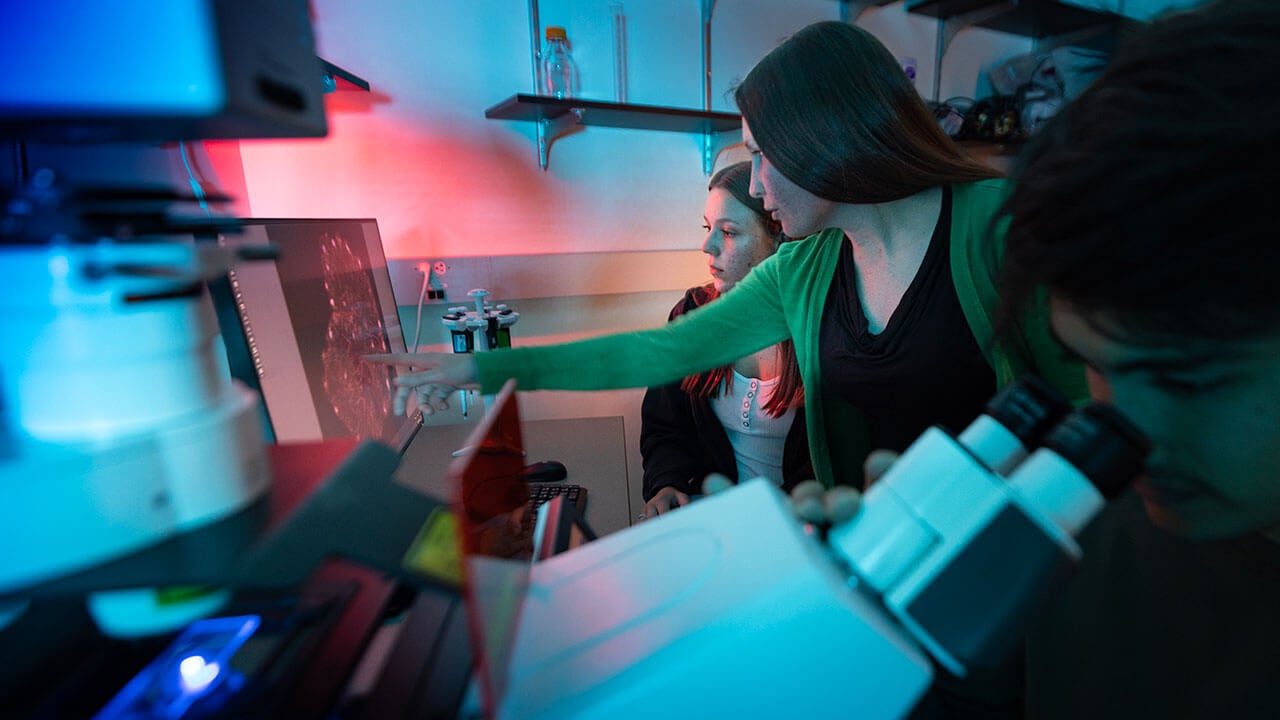Professor Caitlin Hanlon peers over the shoulder of a student in a biology lab and assists them with their microscope