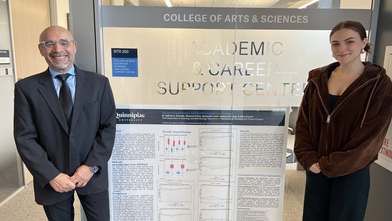 A male professor and female student stand on either side of a research poster showing paragraphs of text, graphs and charts.