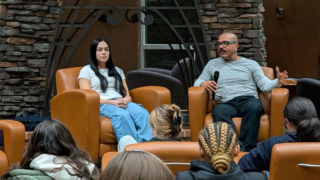 A female and a male speaker sit in chairs in the Piazza; the man holds a microphone.