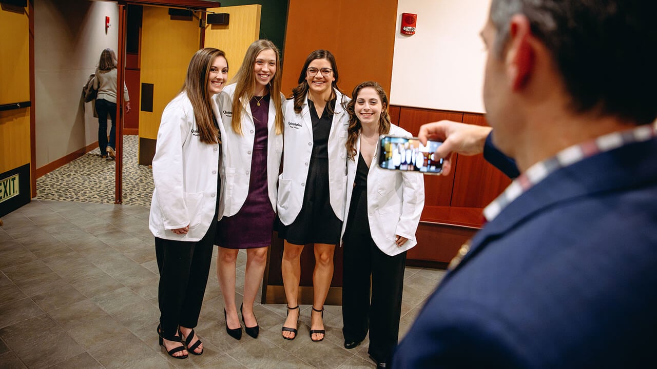 Four DPT white coat students standing and smiling as someone takes their photo in the foreground