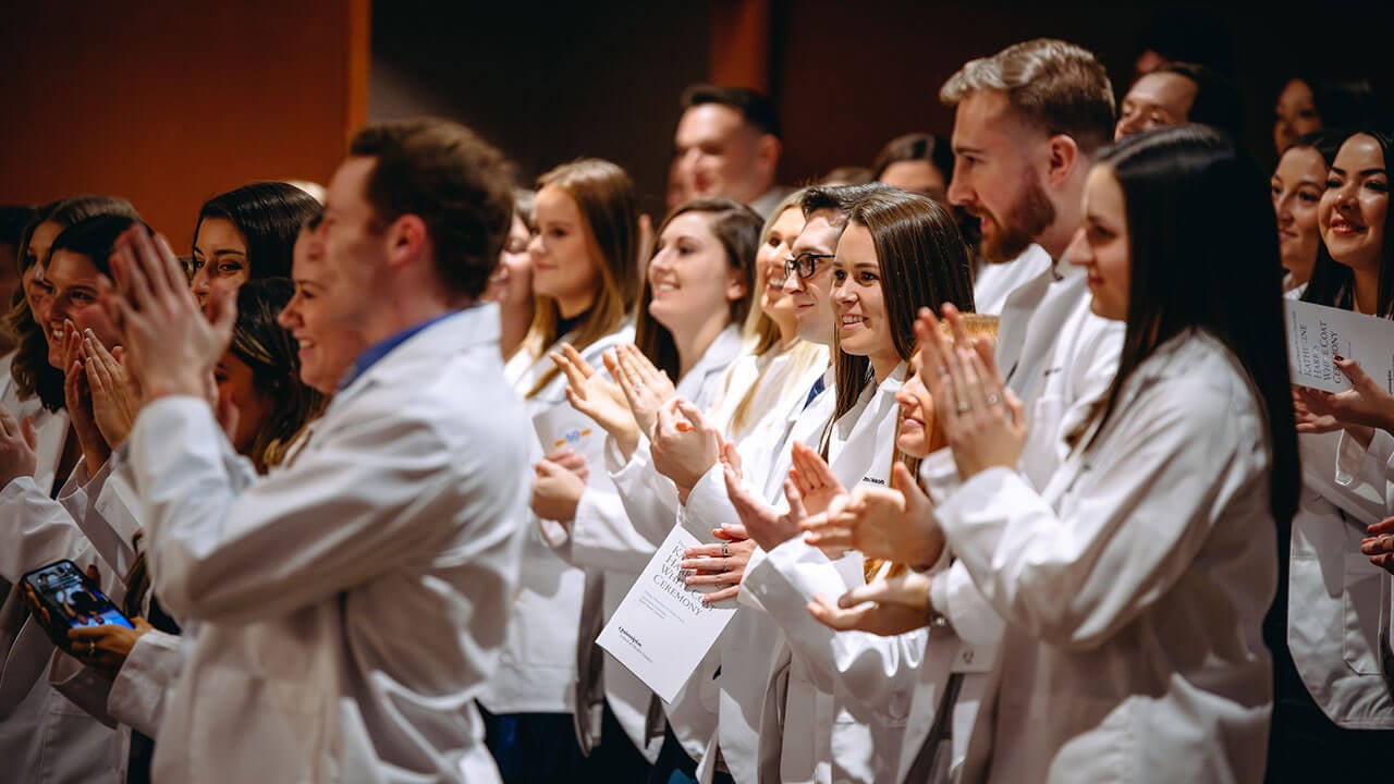 A group of white coat students standing, smiling and clapping together.