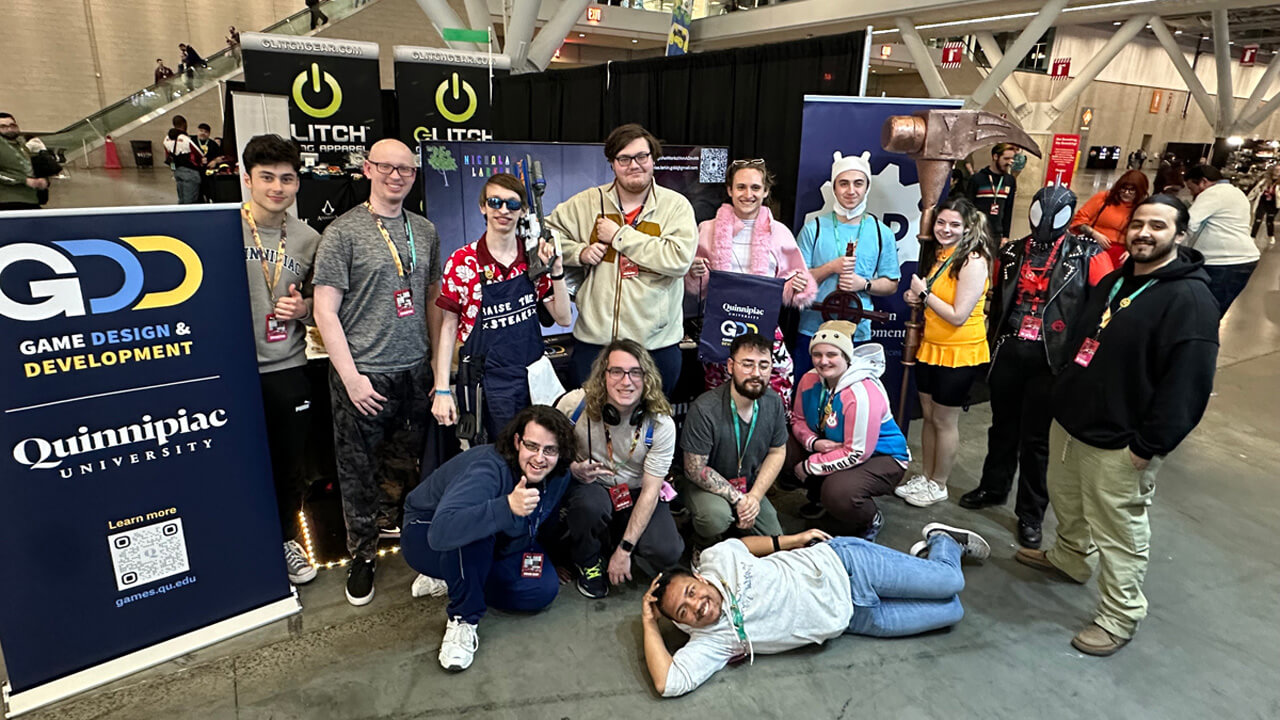 A group of 15 students, some in cosplay costumes, stand in the event hall next to a standing Quinnipiac GDD banner.